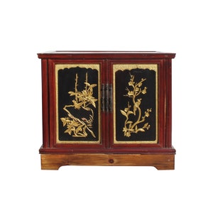 May include: A red wooden cabinet with two doors. The doors are decorated with black panels and gold floral designs. The cabinet has a wooden base.
