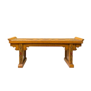 Puede incluir: Un banco de madera con intrincados diseños florales pintados en rojo, amarillo y verde. El banco tiene una parte superior rectangular y dos patas a cada lado. El banco está hecho de madera marrón claro.