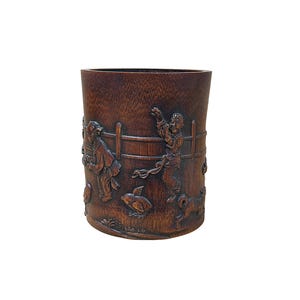 May include: A cylindrical, dark brown wooden container with carved relief figures. The figures depict people, animals, and a fence. The container has a smooth, polished exterior and a dark interior. The overall theme is a rustic, handcrafted aesthetic.