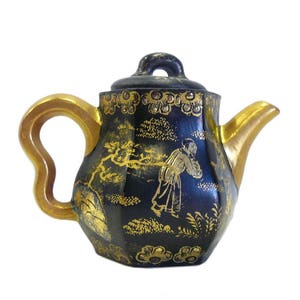 May include: A small, black teapot with gold accents and a floral design. The teapot has a rounded body and a short spout. The handle is gold and has a unique shape.