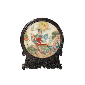 May include: A round, wooden, decorative stand with a carved design. The stand holds a circular, painted panel depicting a woman in a red and green robe holding a lantern, flying through the clouds with a rabbit. The panel has Chinese characters written on it.