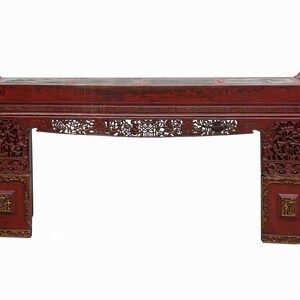 May include: A red wooden table with intricate carvings and gold accents. The table has a rectangular top and two legs on each side. The table is made of wood and has a distressed finish.