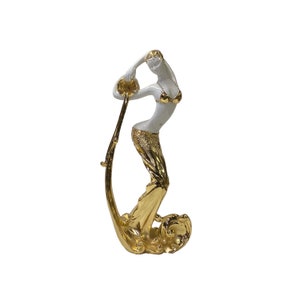 May include: A white figurine of a woman pouring water from a golden pitcher. The woman is wearing a gold skirt and has a gold headband. The figurine is standing on a golden wave.