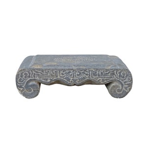May include: A gray stone pedestal with intricate carvings. The top of the pedestal is flat and rectangular, while the base is curved and features a scroll-like design.
