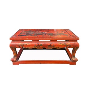 Puede incluir: Una mesa de centro de madera roja con intrincados diseños pintados en negro y dorado. La mesa tiene una tapa rectangular y cuatro patas con pies curvos.