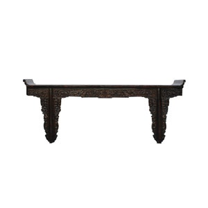 May include: A dark brown wooden console table featuring an elongated rectangular top with upturned ends. The apron and the two sturdy legs are decorated with intricate, deep relief carvings of floral and scroll motifs throughout the structure.