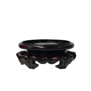 May include: A dark brown wooden pedestal with a round top and three intricately carved legs. The pedestal is designed to display objects.