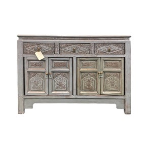 May include: A gray wooden cabinet with intricate carvings and brass hardware. The cabinet has three drawers and four doors.