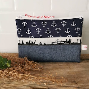Könnte beinhalten: Eine blaue und weiße Reißverschlusstasche mit einem Jeansboden. Die Tasche hat ein weißes Ankermuster auf blauem Grund. Der mittlere Teil zeigt eine schwarze Silhouette der Hamburger Skyline mit dem Wort "Hamburg" darunter.