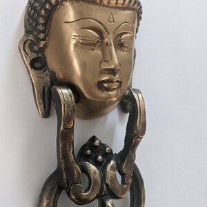 May include: A brass door knocker featuring a detailed Buddha head with a traditional headdress. The knocker has a decorative ring and intricate floral designs.