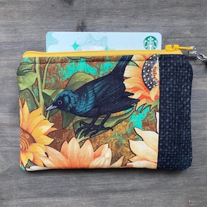 May include: A small, zipped coin purse featuring a bird and sunflower design. The purse has a yellow zip and a black beaded charm. A card is partially visible inside the purse.
