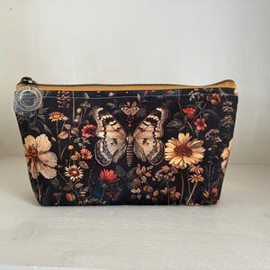 May include: A zipped pouch featuring a black background with a detailed butterfly and various orange, yellow, and red wildflowers. A circular tag attached to the zip pull reads Limited Edition. The pouch has a mustard yellow zip lining.