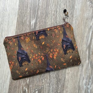 May include: A rectangular zipped pouch featuring a brown fabric with a repeating pattern of hanging bats and scattered autumn leaves in shades of orange, yellow, and brown. The zip pull is decorated with a small black and silver bead charm.