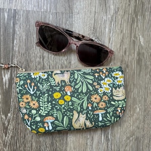 Peut inclure: Une pochette zippée à fond vert foncé ornée d'un motif répétitif de chats, de champignons et de fleurs jaunes et orange, accompagnée d'une paire de lunettes de soleil à monture rose translucide avec des verres foncés et une petite breloque champignon.