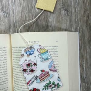 May include: A fabric bookmark with a tea party design, featuring teacups, teapots, and cake, with a yellow felt tea bag attached by a string. The bookmark is placed in an open book.