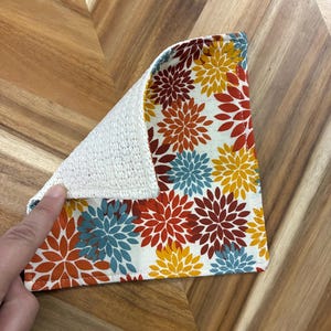 May include: A square cloth with a floral pattern featuring stylised chrysanthemum-like flowers in shades of orange, yellow, and blue on a white background. The reverse side of the cloth has a textured, off-white, woven fabric surface.
