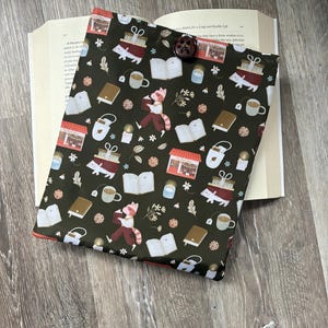 Peut inclure: Une housse de livre en tissu vert foncé avec un motif répétitif de pandas roux lisant des livres, des livres ouverts, des tasses de thé, des bougies et des petites devantures de magasins. La housse est fermée par un bouton marron sur un livre.