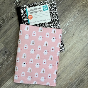May include: A pink fabric notebook cover featuring a repeating pattern of small white ghosts, layered over a black and white speckled composition book labelled Pen+Gear with 200 pages and dimensions of 19 cm x 24.8 cm.