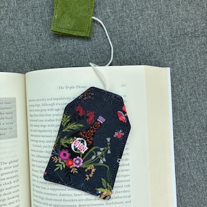 May include: A fabric bookmark shaped like a tea bag with a dark navy background featuring a floral print and a bottle labelled POISON. A white string connects the bookmark to a small, square, green felt tag resting above the open book pages.