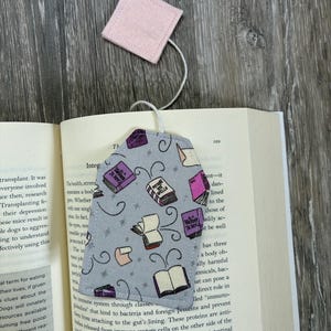 May include: A fabric bookmark shaped like a tea bag with a light grey background featuring a pattern of colourful books and scrolls. A white string connects the bookmark to a small, square, light pink fabric tag resting above the open book.
