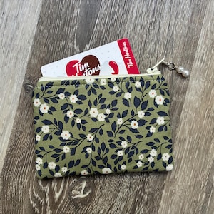 May include: A rectangular fabric coin purse featuring an olive green background with a repeating pattern of dark navy leaves and small white flowers with yellow centres. A white zip closure includes a small charm with two white faux pearls.