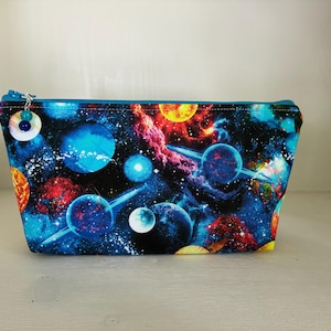 Peut inclure: Une trousse rectangulaire à fermeture éclair avec un imprimé spatial vibrant représentant des planètes, des étoiles et des nébuleuses dans des tons de bleu, rouge, orange et jaune. La tirette est ornée d'une breloque argentée avec deux perles.
