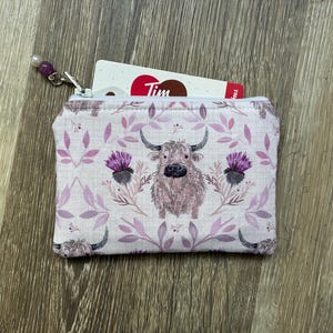 May include: A rectangular fabric coin purse featuring a repeating pattern of Highland cattle and purple thistle flowers on a light background. The zip pull is decorated with a purple and white bead. A white card with Tim text is inside.