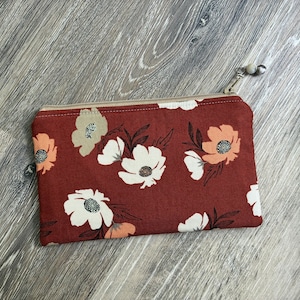 May include: A rectangular fabric zip pouch in a deep rust-red colour featuring a floral pattern with white, peach, and beige flowers and dark brown leaves. The pouch has a beige zip closure with a small silver-toned metal bead charm attached.