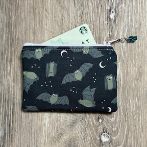 May include: A rectangular fabric zip pouch featuring a dark navy blue background with a repeating pattern of light green bats, white crescent moons, and small white stars. A white card with a green logo is partially inserted into the top opening.
