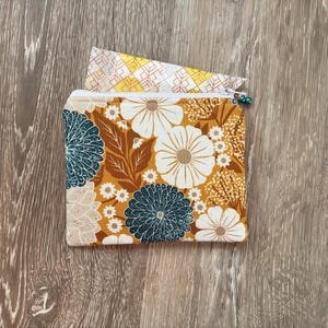 Peut inclure: Une pochette zippée rectangulaire en tissu avec un fond jaune moutarde orné d'un motif floral de fleurs et de feuilles blanches, bleu sarcelle foncé et beige. Un morceau de tissu à motif géométrique jaune et blanc dépasse du haut.