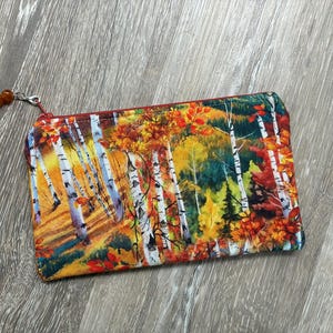 Peut inclure: Une pochette rectangulaire à fermeture éclair avec un motif de forêt imprimé sur tissu, représentant des bouleaux blancs et un feuillage d'automne dans des tons jaune, orange et rouge. La pochette possède une fermeture rouge avec deux perles.