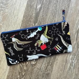 Peut inclure: Une trousse rectangulaire en tissu à fond noir ornée d'un motif répétitif de dinosaures en blouse de laboratoire, de formules chimiques et d'équipement scientifique. La trousse possède une fermeture éclair bleu vif avec une perle.