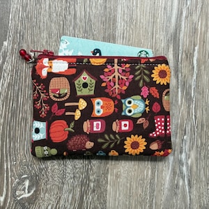 Peut inclure: Une pochette zippée en tissu rectangulaire avec un fond marron foncé présentant un motif automnal répétitif d'hiboux, de renards, de citrouilles, de tournesols, de nichoirs, de champignons et de bottes rouges à pois. Une carte bleu clair est insérée.