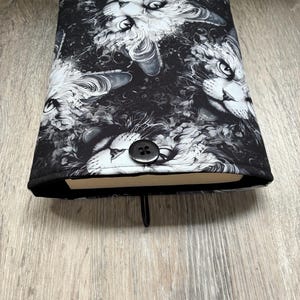 Peut inclure: Une housse de livre en tissu avec un motif répétitif en noir et blanc représentant des visages de chats aux textures de fourrure détaillées. La housse rectangulaire est fermée par un bouton noir et possède une boucle élastique noire en bas.