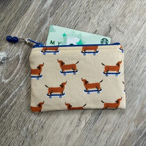 Peut inclure: Une pochette zippée rectangulaire en tissu beige avec un motif répétitif de teckels marron sur des skateboards bleus. La pochette possède une fermeture éclair bleue avec deux petites perles bleues sur la tirette. Une carte verte est insérée.