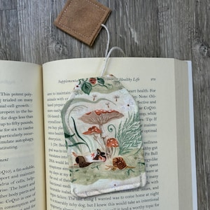 May include: A fabric bookmark shaped like a tea bag with a printed pattern of mushrooms, a snail, and a small animal. The bookmark features a cream-coloured background with green foliage and is attached to a small, square, brown felt tag by a white string.
