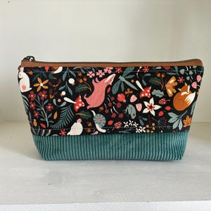 May include: A rectangular cosmetic bag with a black background featuring a colorful woodland pattern of foxes, mushrooms, and flowers. The bottom half is teal corduroy, and the zipper is brown.