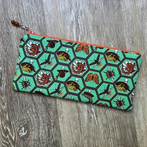 Peut inclure: Une trousse rectangulaire à fermeture éclair avec un motif répétitif d'hexagones noirs sur fond vert menthe. À l'intérieur des hexagones se trouvent des illustrations d'insectes variés, dont des papillons, des abeilles et des coléoptères.