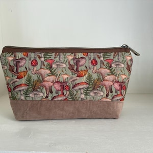 Peut inclure: Une trousse zippée avec une partie supérieure en tissu vert clair ornée d'un motif répétitif de champignons et de fougères, et une partie inférieure en velours côtelé marron uni. La trousse possède une fermeture éclair marron sur le dessus.