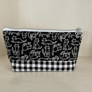 May include: A zipped pouch featuring a black fabric upper section with a white line-art cat pattern and a black and white gingham checkered fabric lower section. The silver zip pull is shaped like a small metal paw print charm.
