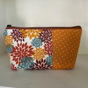 Peut inclure: Une pochette zippée présentant un fond blanc avec un motif floral de fleurs ressemblant à des dahlias rouges, orange et bleus sur la gauche, et un tissu jaune moutarde uni avec un motif de croix blanches sur la droite.