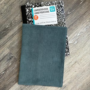 May include: A teal corduroy notebook cover placed over a black and white speckled composition book. The composition book label reads COMPOSITION BOOK, 200, and 19 cm x 24.8 cm. The cover features vertical ribbed texture and stitched edges.