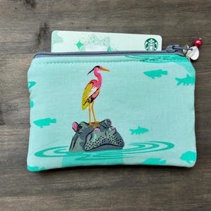 May include: A small, zipped pouch with a teal background featuring a colourful bird standing on a hippo in water, with fish and water ripples. A Starbucks gift card is partially visible. The zipper pull has a red bead.