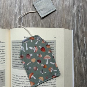 May include: A fabric bookmark shaped like a tea bag with a sage green background featuring a repeating pattern of various mushrooms in shades of brown, orange, and white, connected by a white string to a small, square, light grey felt tab.
