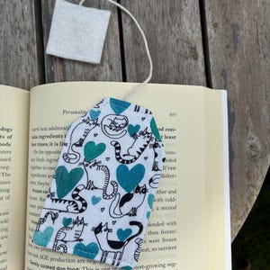 May include: A fabric bookmark shaped like a tea bag with a white background featuring a pattern of black and white cats and teal hearts. A white string is attached to the top, leading to a small, square, white felt tag at the end of the string.