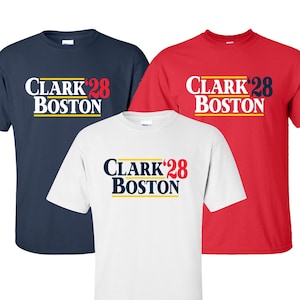 May include: Three t-shirts in navy blue, red, and white. Each shirt features the text "CLARK '28 BOSTON" in white, red, and navy blue, respectively, with a yellow stripe beneath the text. The shirts are short-sleeved.