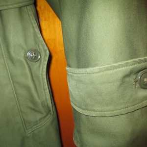 May include: Close-up of a green military-style jacket with a buttoned flap pocket and a buttoned cuff. The fabric has a textured, slightly worn look.
