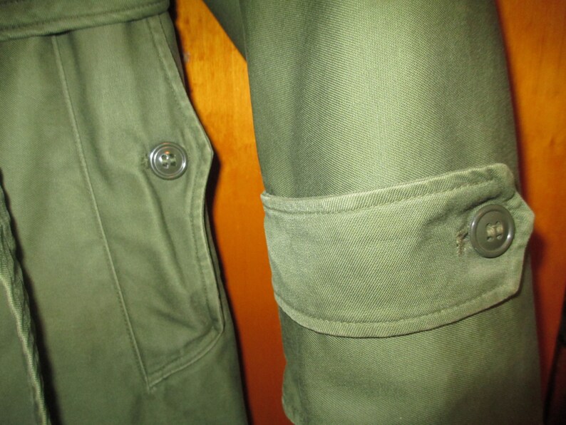May include: Close-up of a green military-style jacket with a buttoned flap pocket and a buttoned cuff. The fabric has a textured, slightly worn look.