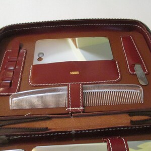 May include: Brown leather travel kit with a zipper closure. The kit is open and contains a mirror, a comb, a nail file, and a small container with multiple compartments.