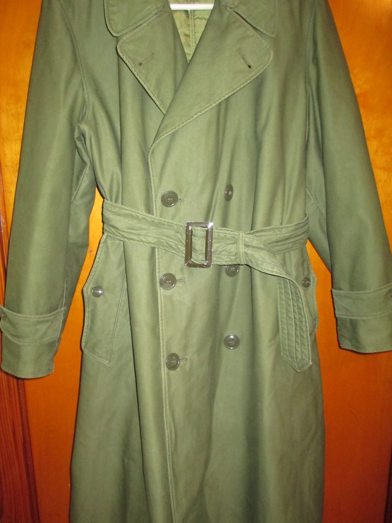 May include: A green double-breasted trench coat with a belt and four buttons. The coat has two pockets on the front and a single pocket on the left sleeve.